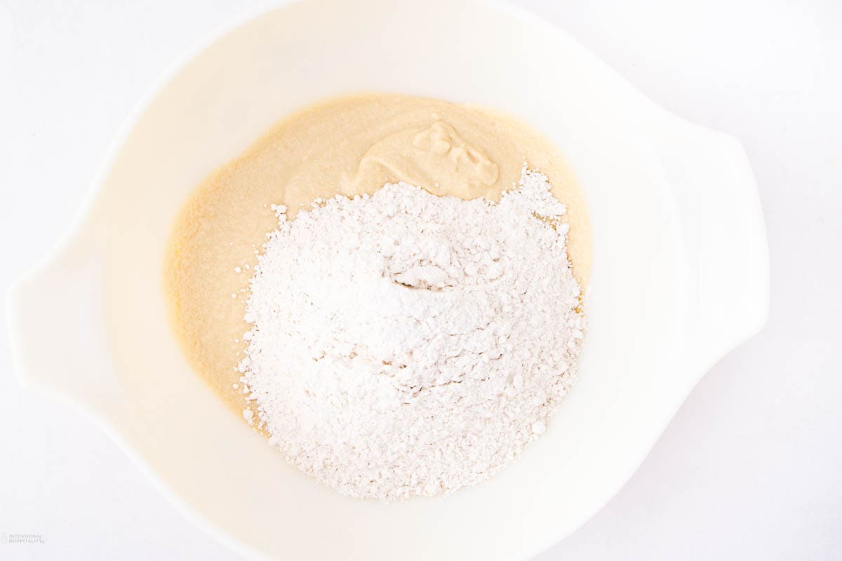 A white mixing bowl contains flour and a wet batter mixture, ready to be stirred together. The ingredients contrast in texture and color, with the flour sitting atop the smooth, slightly tan batter. The background is white.