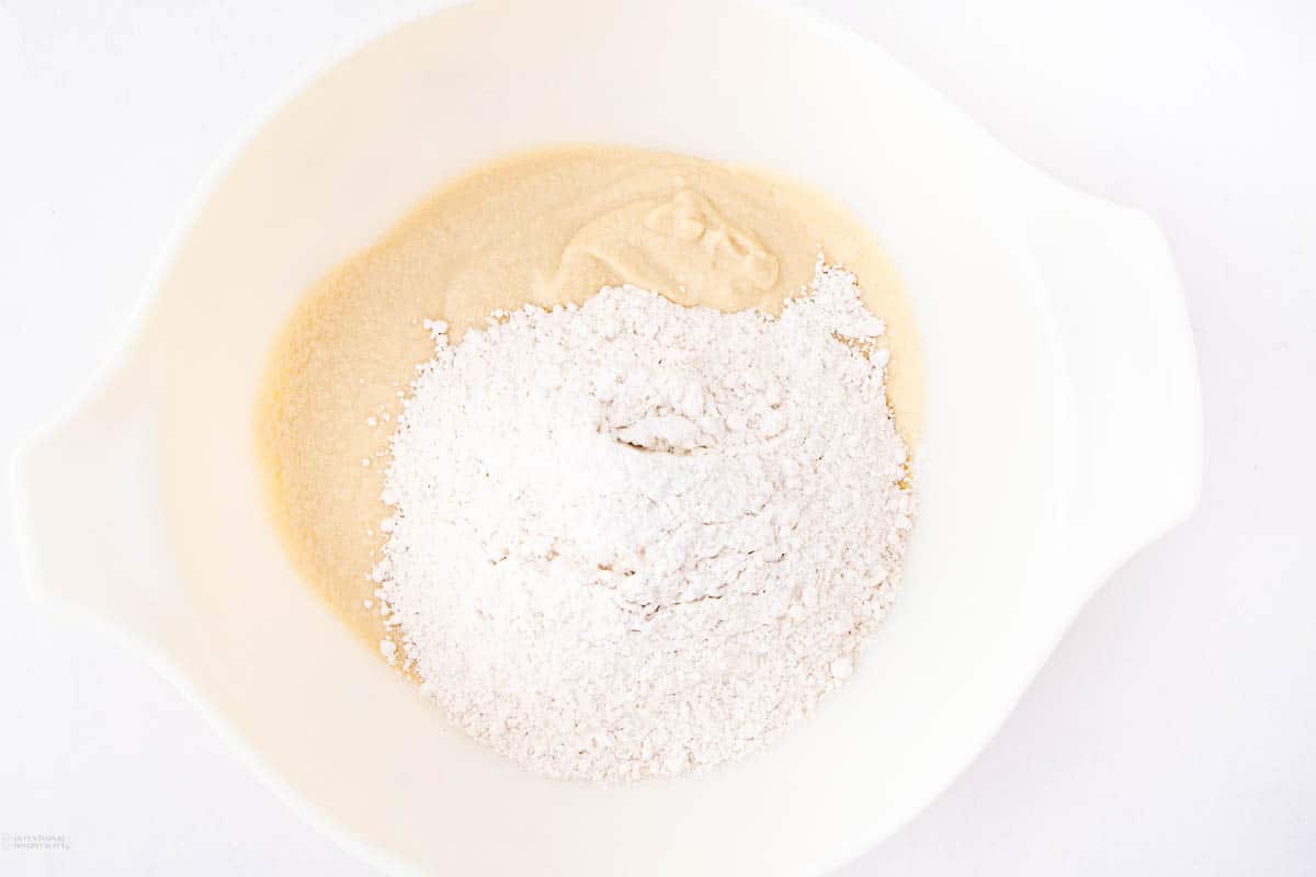 A white mixing bowl containing a pale batter and a mound of flour on one side. The ingredients are about to be mixed together on a white surface.