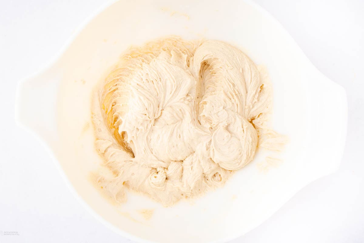 A white bowl filled with light beige whipped cream or frosting, showing a smooth and creamy texture. The background is plain and white, emphasizing the contents of the bowl.