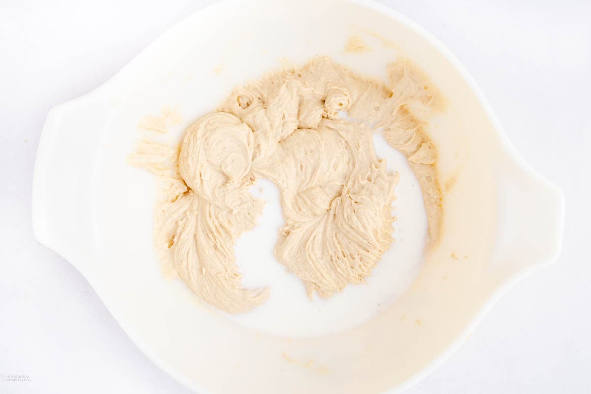 A white mixing bowl containing creamy beige dough, partially blended with a white liquid, possibly milk. The mixture has a smooth texture with small air pockets visible. The bowl is placed on a light surface.