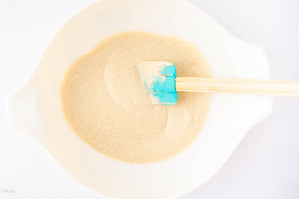 A white bowl containing creamy batter is being mixed with a light blue silicone spatula with a wooden handle. The bowl is set against a plain white background.