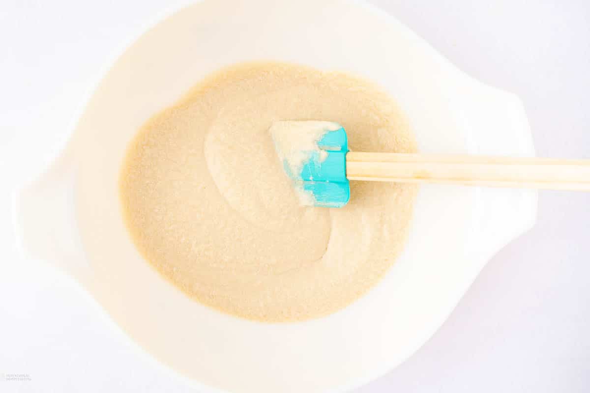 A white mixing bowl containing a pale batter is being stirred with a teal spatula with a wooden handle. The image is taken from above, showing the smooth, creamy texture of the batter.