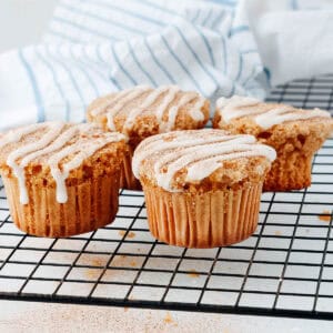 Four cinnamon spice muffins with white icing drizzle sit on a black cooling rack. In the background, a white and blue striped cloth is partially visible. The muffins are golden brown, highlighting their freshly baked texture.