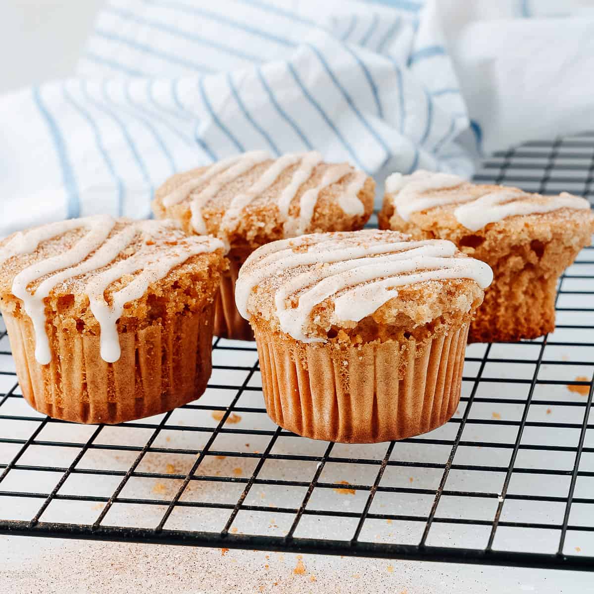 four muffins topped with glaze, sugar, and cinammon on a cooling rack, a dish towel behind