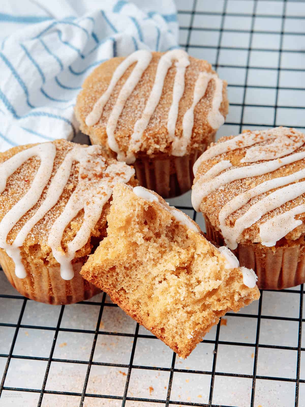 Three cinnamon muffins with icing drizzled on top are placed on a cooling rack. One muffin is torn in half, revealing a soft, moist interior. A blue and white striped cloth is in the background.