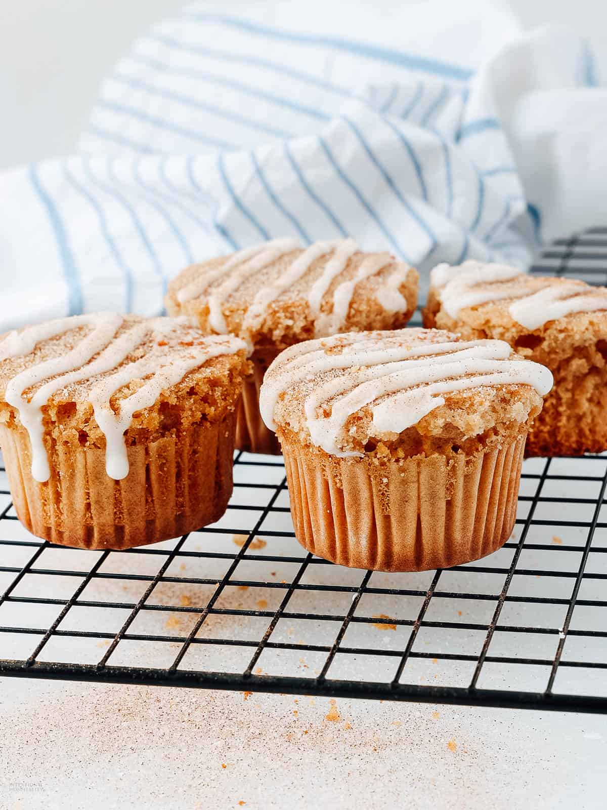 Four muffins drizzled with icing sit on a black cooling rack. The muffins are lightly dusted with cinnamon sugar, and a striped blue and white cloth is in the background.