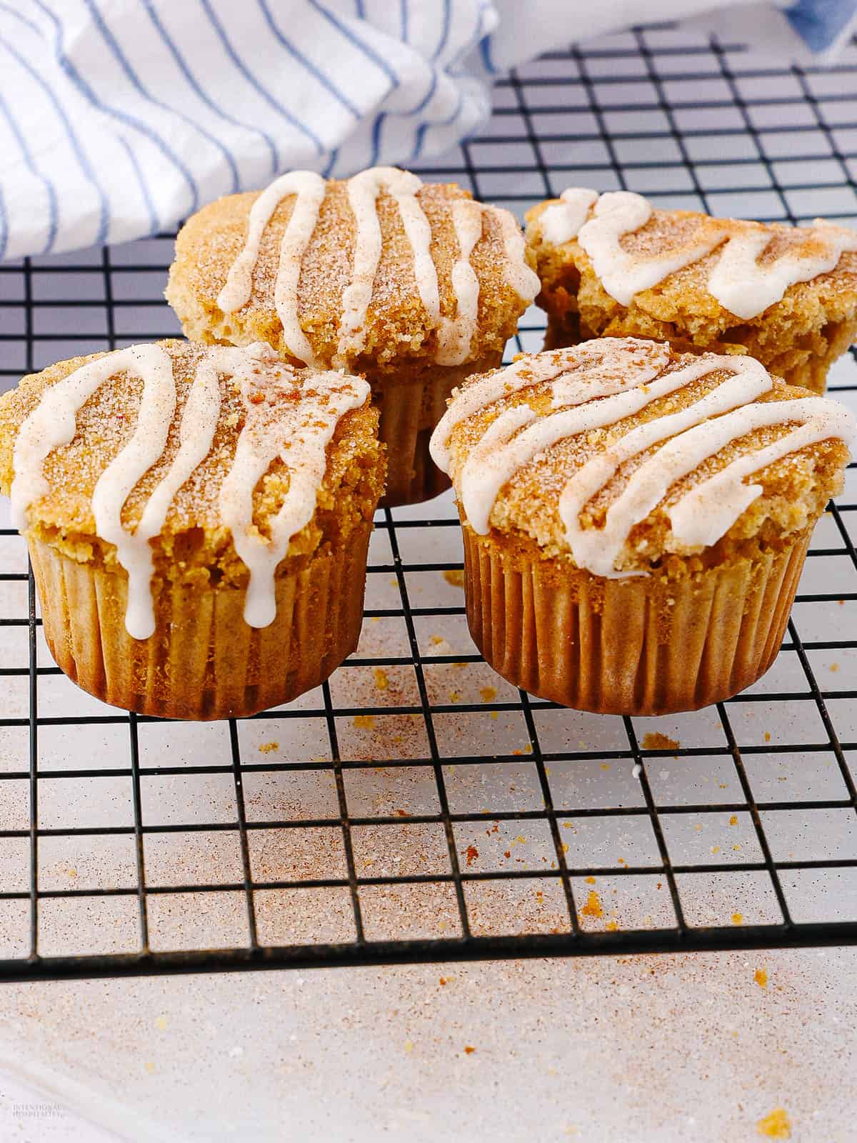 Four cinnamon muffins with white icing drizzle are cooling on a black wire rack. A striped blue and white cloth is partially visible in the background. The muffins are surrounded by a light dusting of cinnamon.