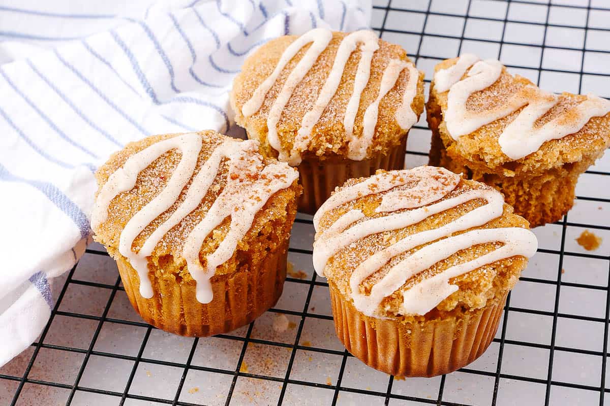 Four cinnamon muffins with white icing drizzle sit on a cooling rack. A striped cloth is partially visible beside them.