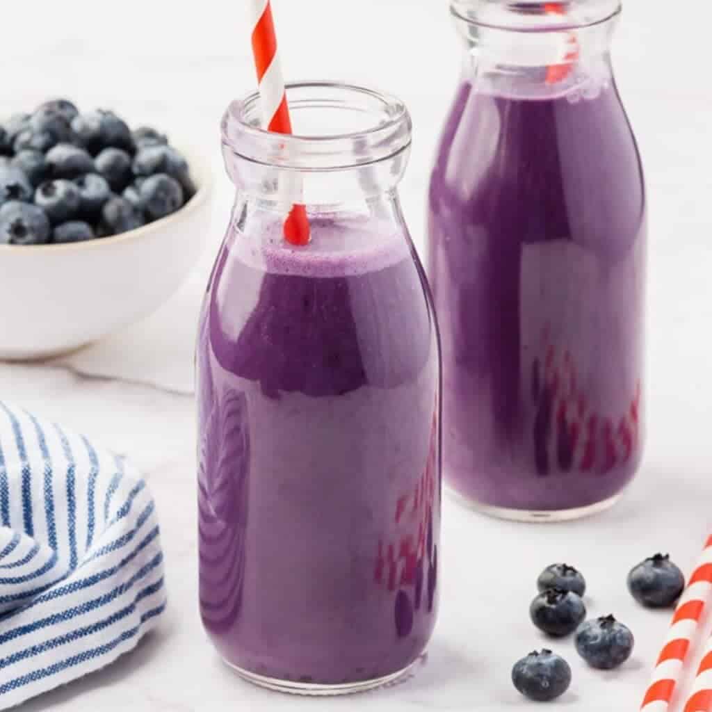 Two glass bottles filled with purple blueberry smoothies, each with a red and white striped straw. In the background, a bowl of fresh blueberries is visible next to a striped cloth and scattered blueberries on a white surface.