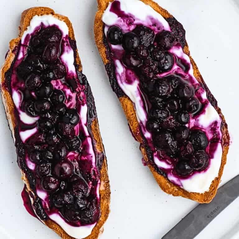 Two slices of toasted bread topped with creamy yogurt and a generous layer of blueberry compote. The compote has a rich, dark purple hue, while the yogurt provides a stark white contrast. The toast is served on a white plate with a knife.