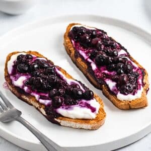 Two slices of toasted bread topped with creamy yogurt and a generous layer of blueberries on a white plate, with a fork beside them.