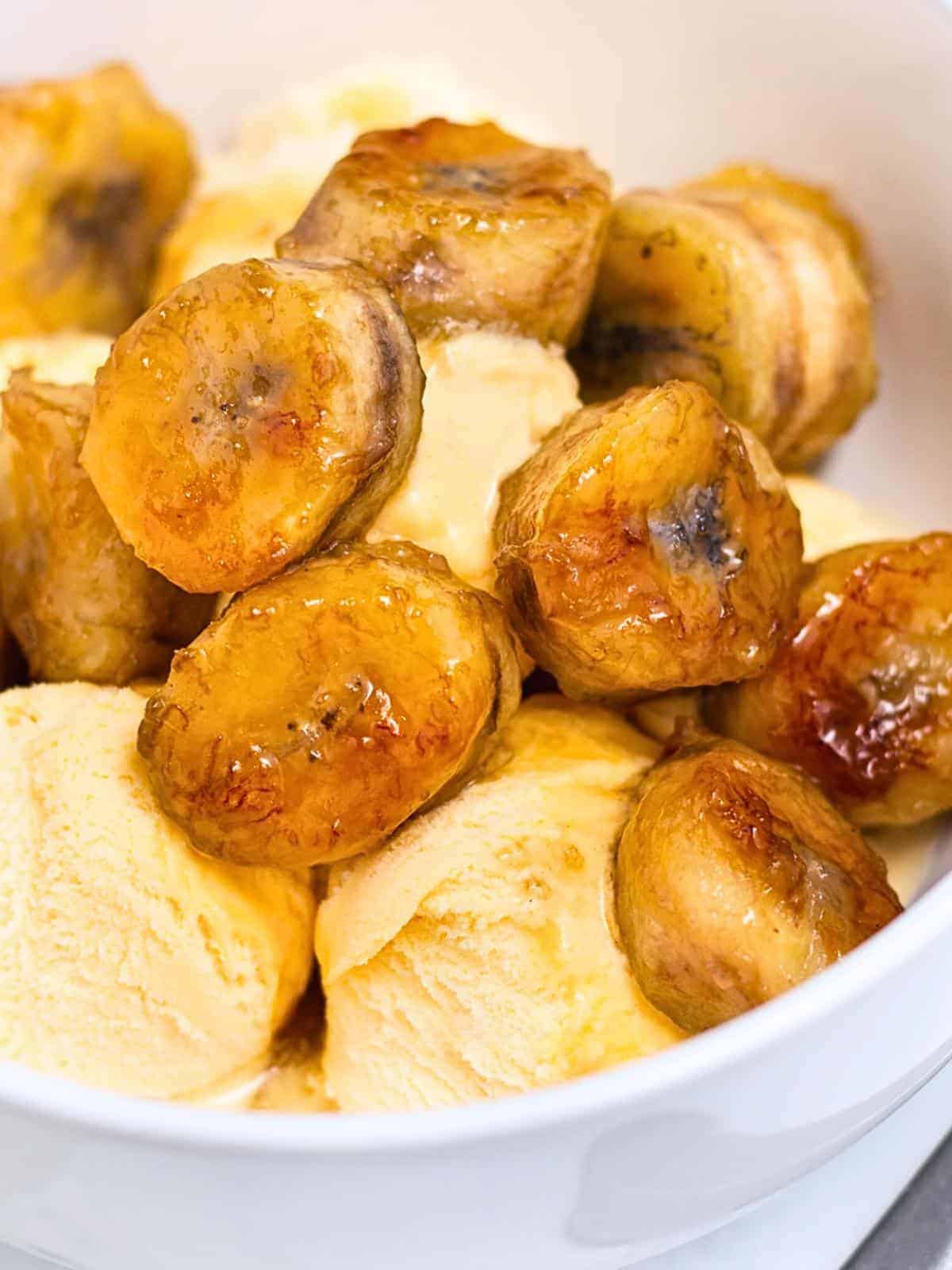 A bowl of caramelized banana slices served atop scoops of vanilla ice cream. The bananas are golden brown, glistening with syrup, and contrast with the creamy texture of the ice cream.