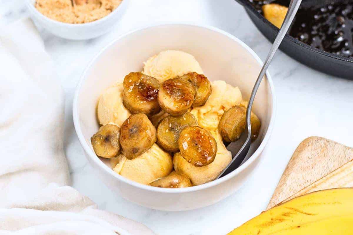 A bowl of vanilla ice cream topped with caramelized banana slices, placed on a white marble surface. A spoon is in the bowl. Nearby, a cutting board with a partially visible banana and a dish of brown sugar are seen.