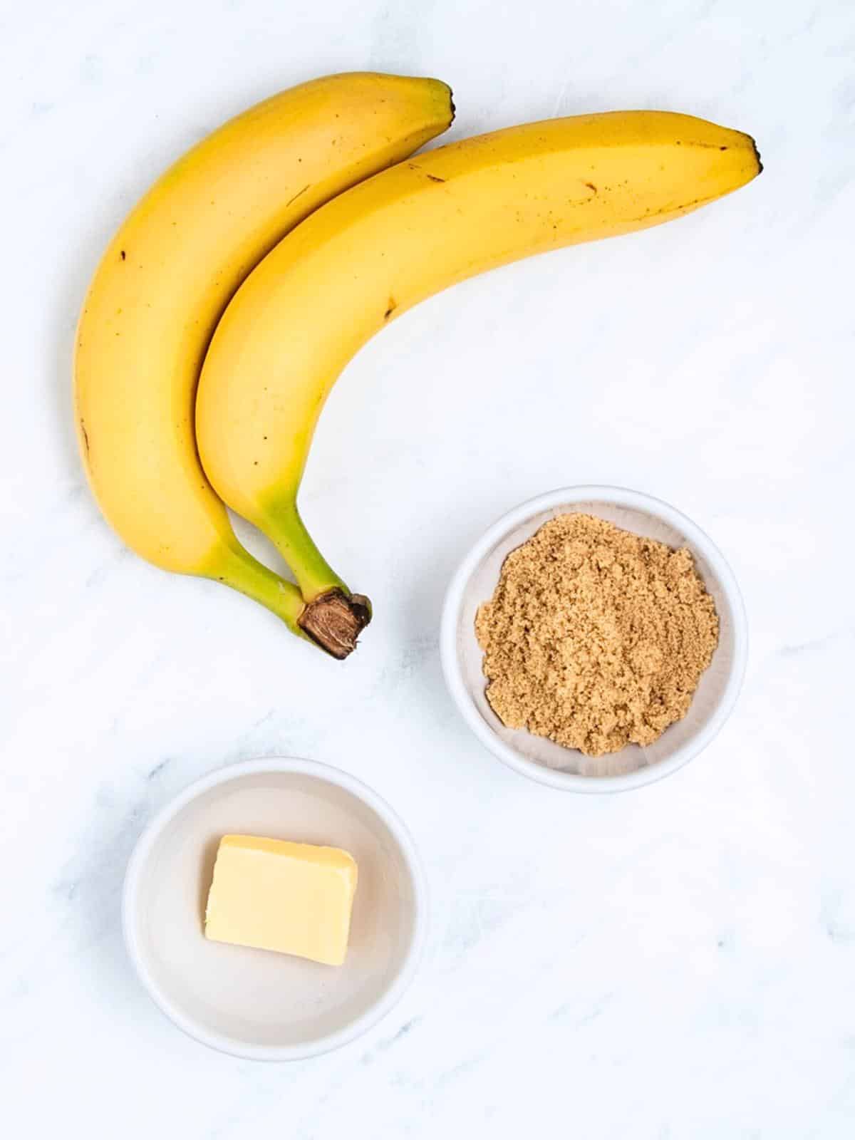 Two yellow bananas, a bowl of brown sugar, and a bowl with a small slab of butter are arranged on a white marble surface.