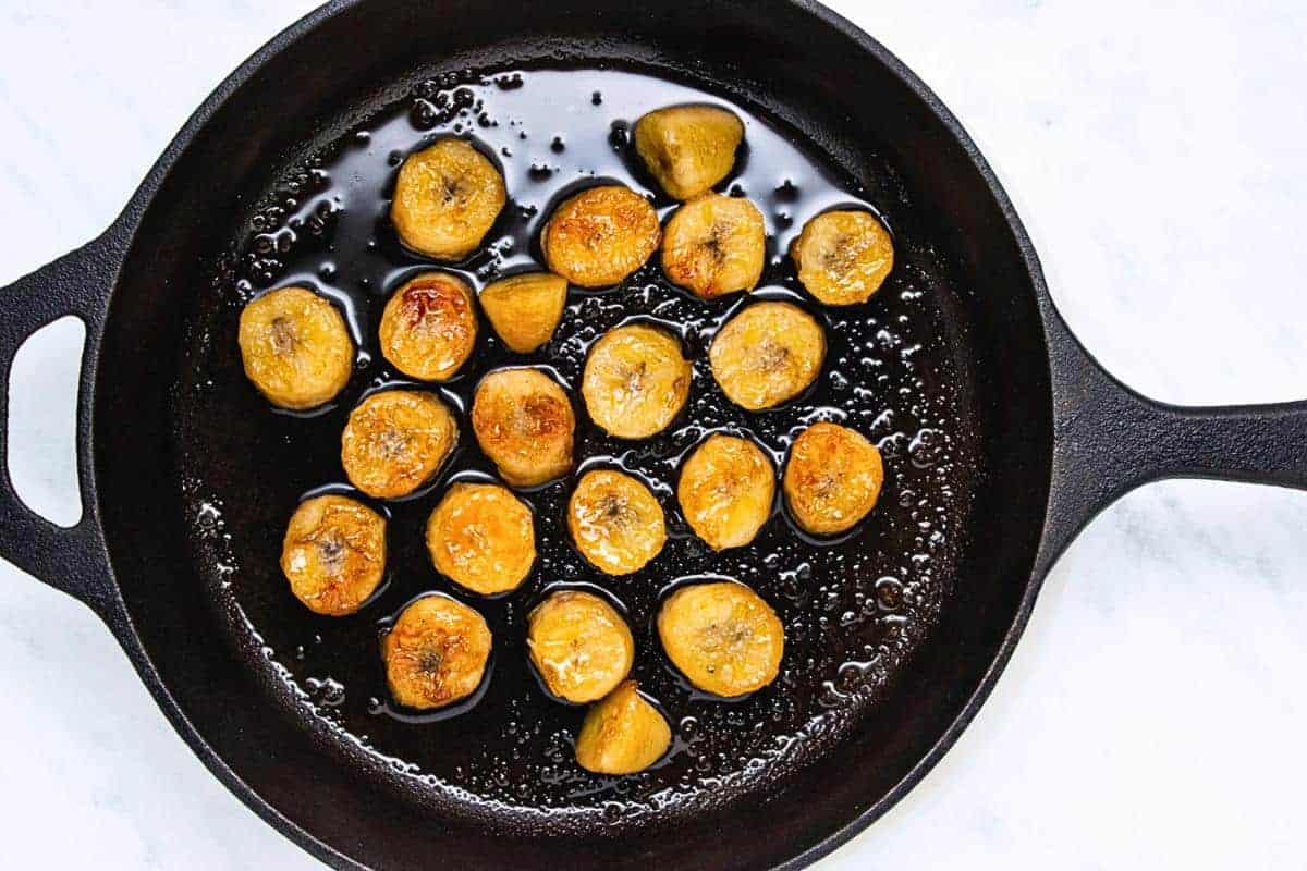 A cast iron skillet with sliced plantains frying in oil. The plantains are golden brown, evenly spaced, and surrounded by bubbling oil. The skillet is placed on a white marble surface.