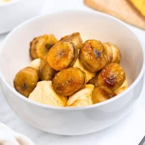 A white bowl filled with scoops of vanilla ice cream topped with caramelized banana slices. The background has a soft focus on other blurred elements.