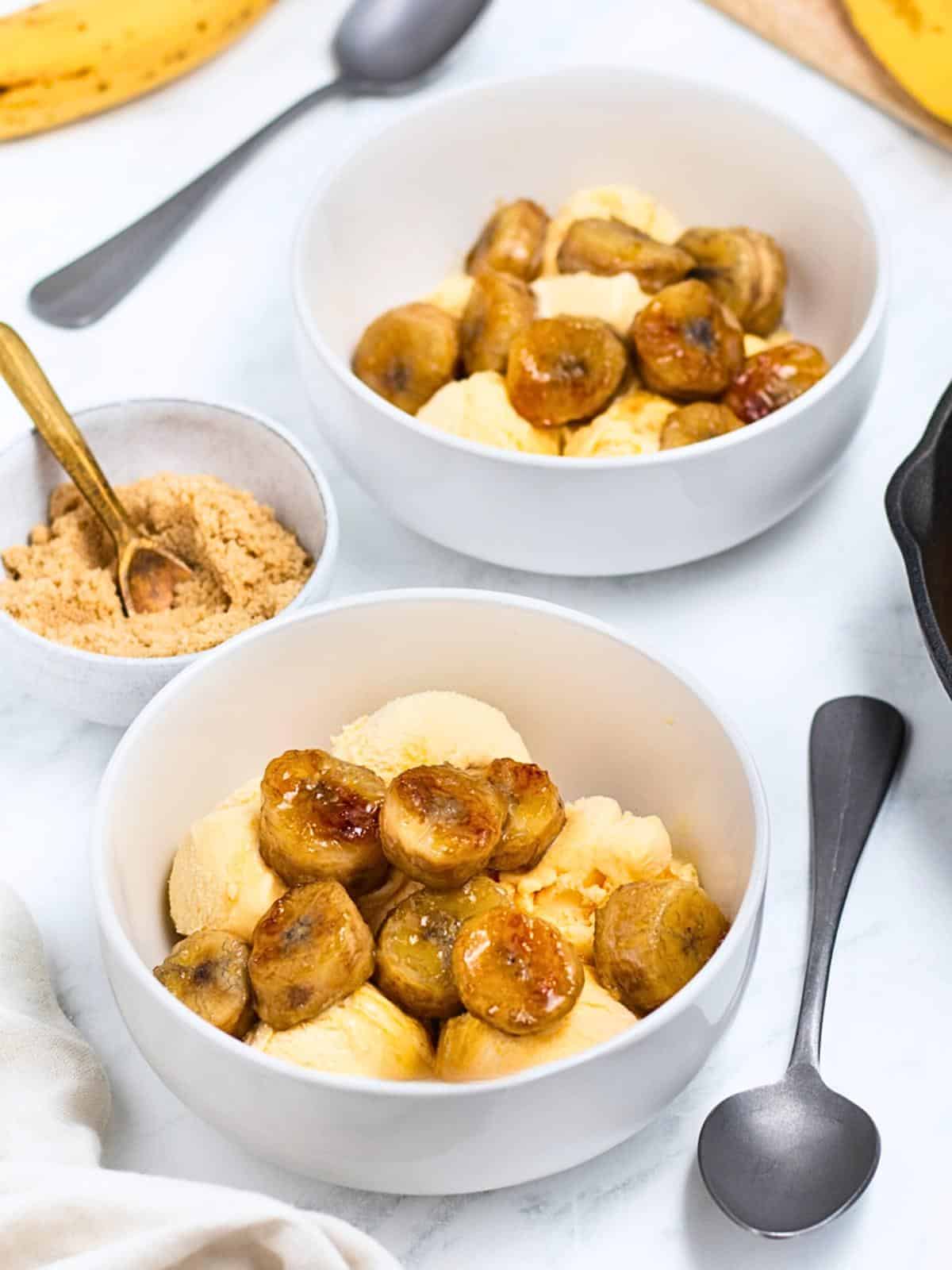 Two bowls of vanilla ice cream topped with caramelized bananas. A small bowl of brown sugar and two spoons are placed nearby on a marble surface. A hint of a banana and skillet are visible in the background.