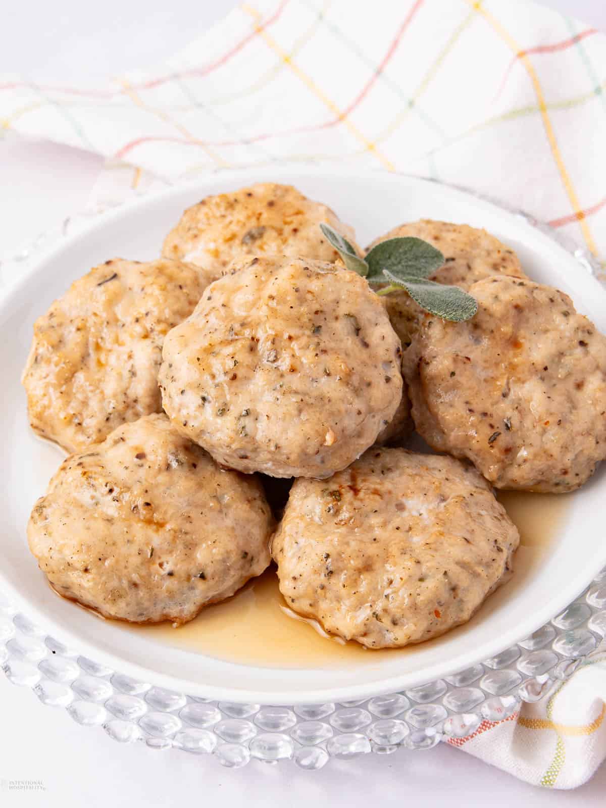 A plate of homemade sausage patties topped with a garnish, served on a textured glass platter. The patties are golden brown, seasoned with herbs, and are sitting on a light, checkered kitchen towel in the background.