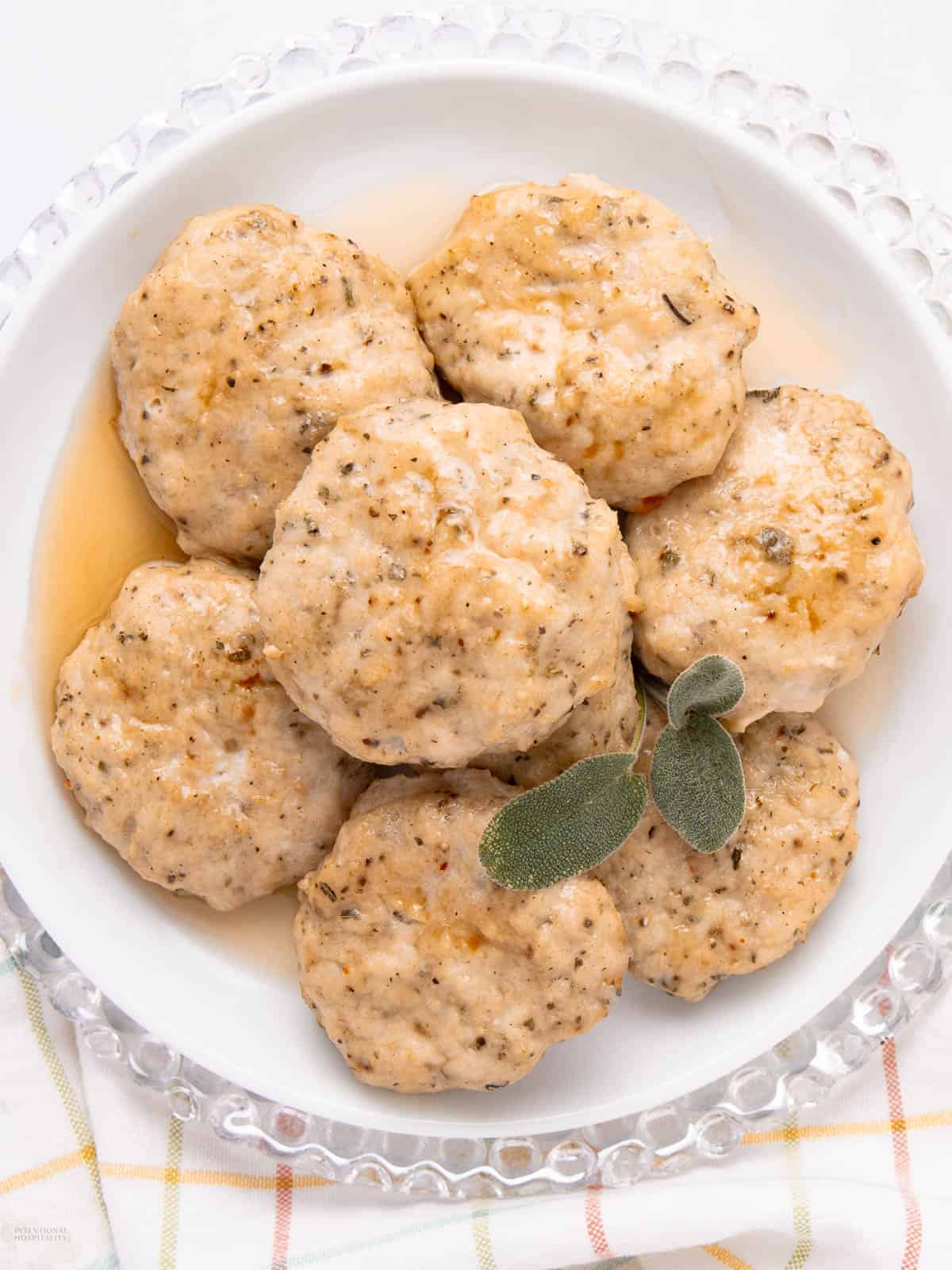 A white round plate with seven sausage patties drizzled with syrup. The patties are seasoned with herbs and spices, and topped with two fresh sage leaves. The plate is on a clear glass charger, placed on a checked cloth.