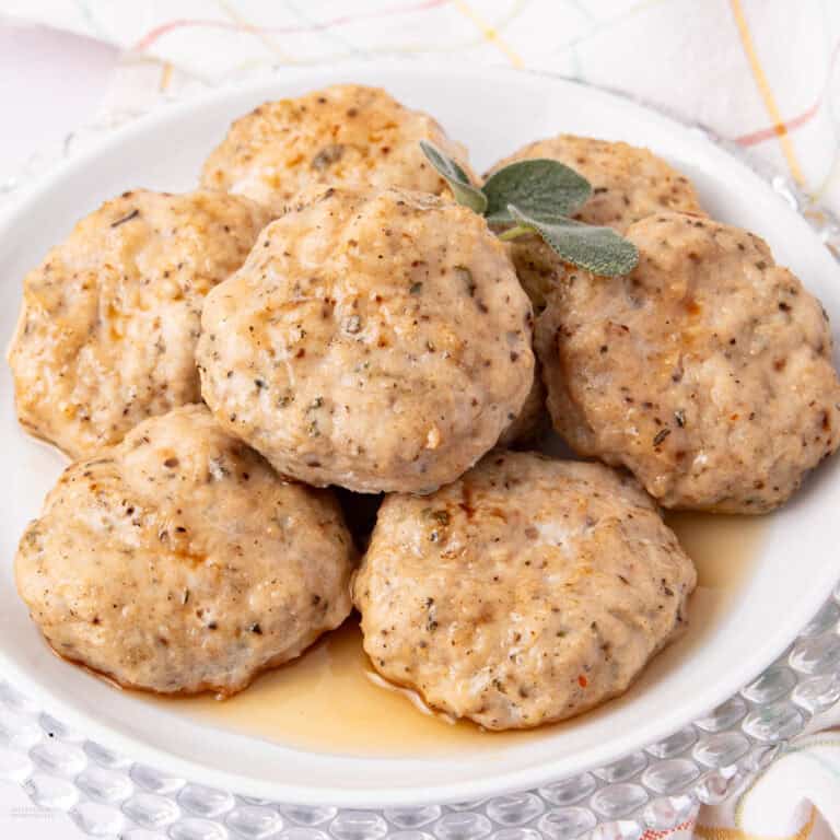 A plate of six browned sausage patties garnished with a sprig of sage. The patties have a seasoned appearance and are served on a white plate with a decorative border. A checkered cloth is faintly visible in the background.