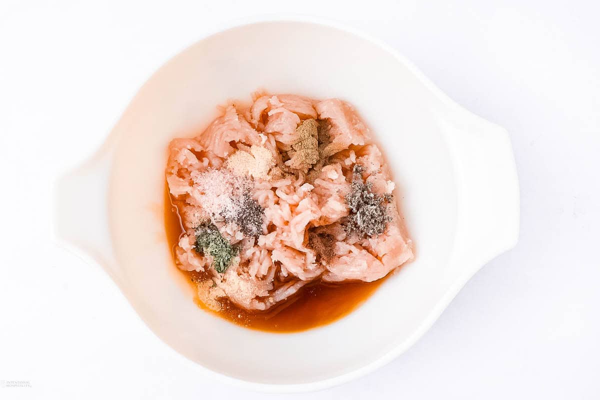 A white bowl contains raw, marinated chicken pieces. The meat is seasoned with various spices, creating a colorful mixture. The background is plain white, emphasizing the marinated chicken in the bowl.