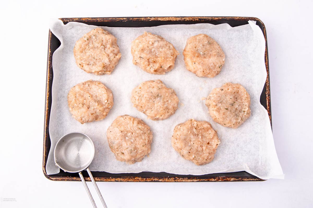 Eight raw, seasoned chicken patties on a baking sheet lined with parchment paper, beside a metal measuring cup.