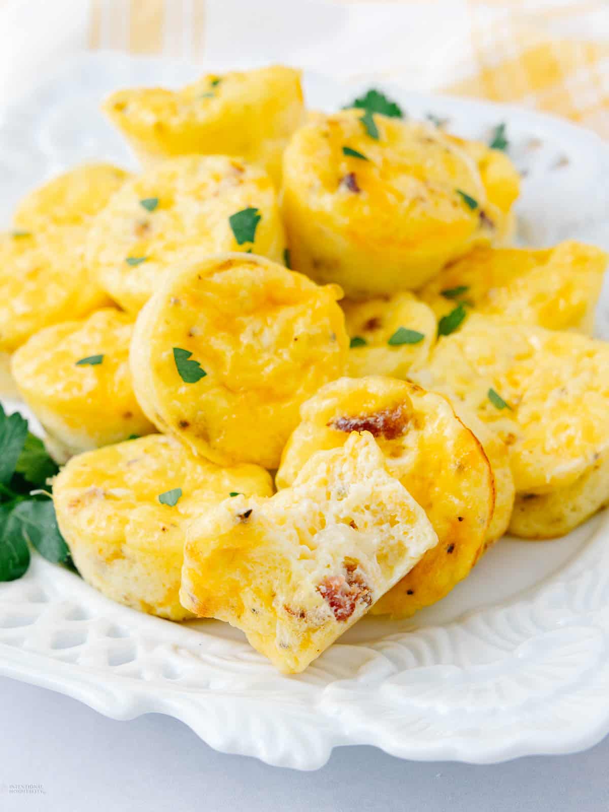 A plate of fluffy, golden egg muffins garnished with chopped herbs. These cottage cheese egg bites, some with visible bits of meat and cheese, are stacked on a decorative white plate. A few sprigs of fresh parsley accompany the arrangement.