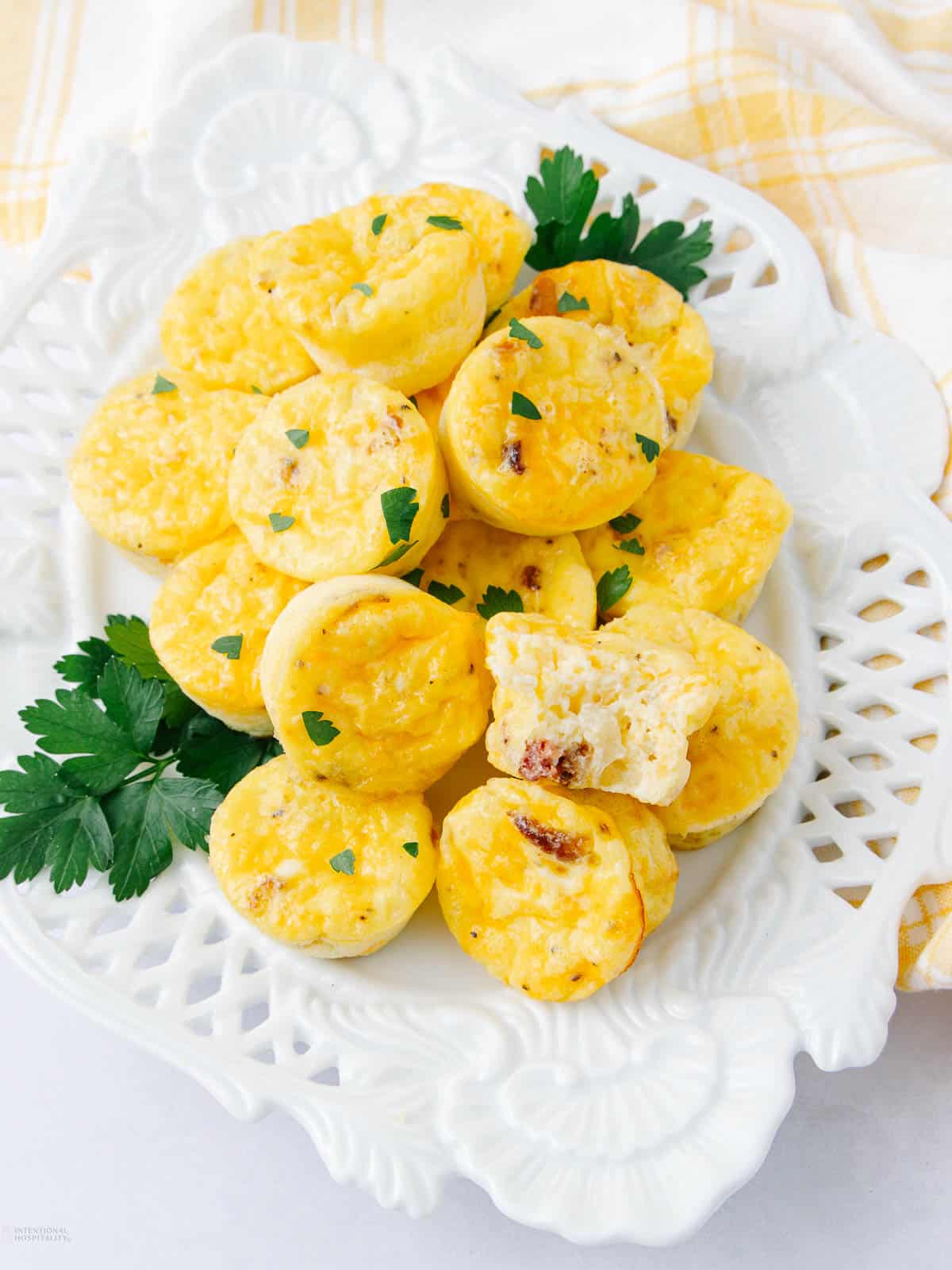 A white plate with several mini egg muffins garnished with parsley. These fluffy cottage cheese egg bites reveal tempting bits of bacon inside. The background is softly accented by a yellow-checkered cloth, enhancing their homey appeal.