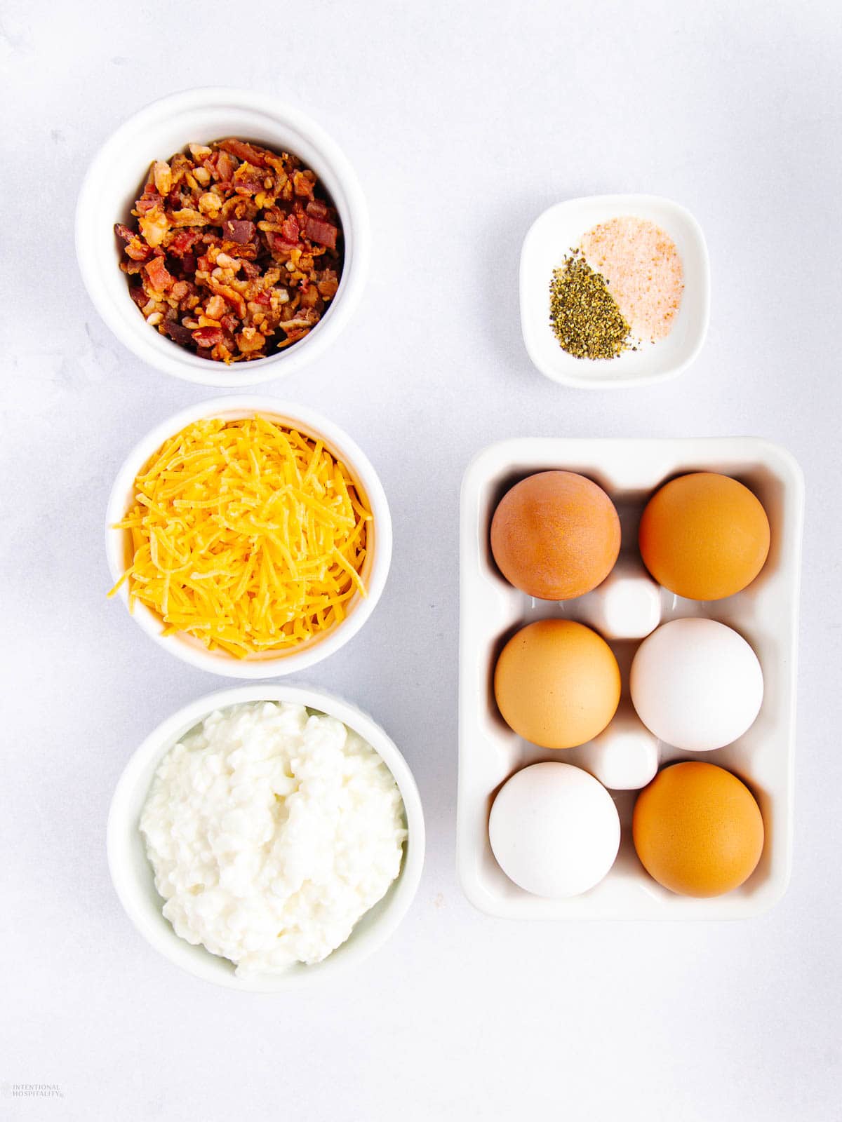 Top-down view of ingredients on a white surface: chopped bacon, shredded cheese, cottage cheese egg bites mix with eggs in a carton, salt and pepper.