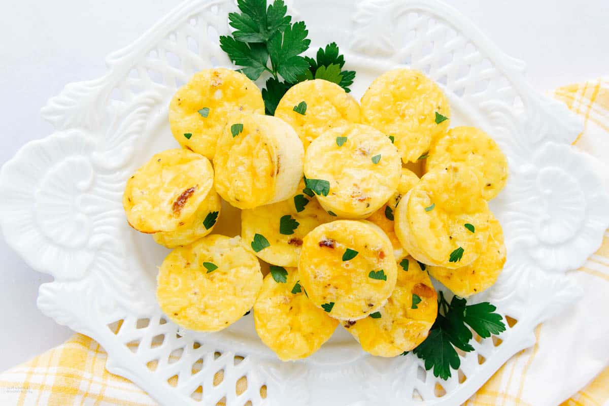 A white plate holds a serving of small, round cottage cheese egg bites, garnished with fresh parsley. The eggs are golden and lightly browned. The plate is placed on a yellow and white checkered cloth.