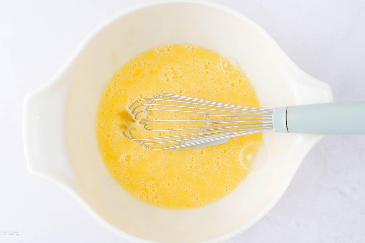 A whisk with a light blue handle is placed in a white bowl containing a beaten yellow mixture, likely the beginnings of cottage cheese egg bites. The bowl rests on a light gray surface.