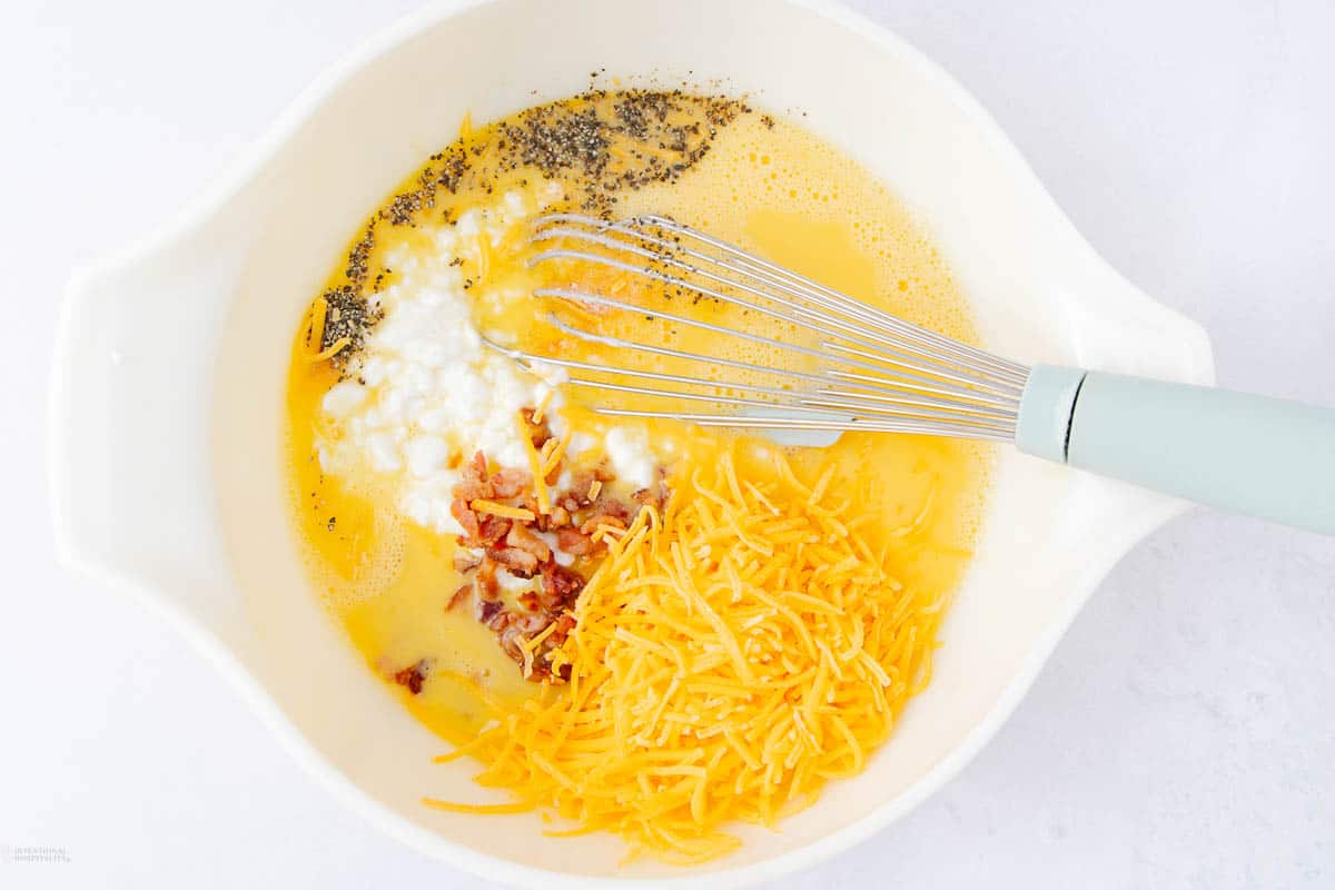 A white bowl holds a savory blend for cottage cheese egg bites, featuring eggs, shredded cheese, bacon bits, and black pepper. A whisk with a green handle sits poised in the bowl, ready to bring these delicious ingredients together.