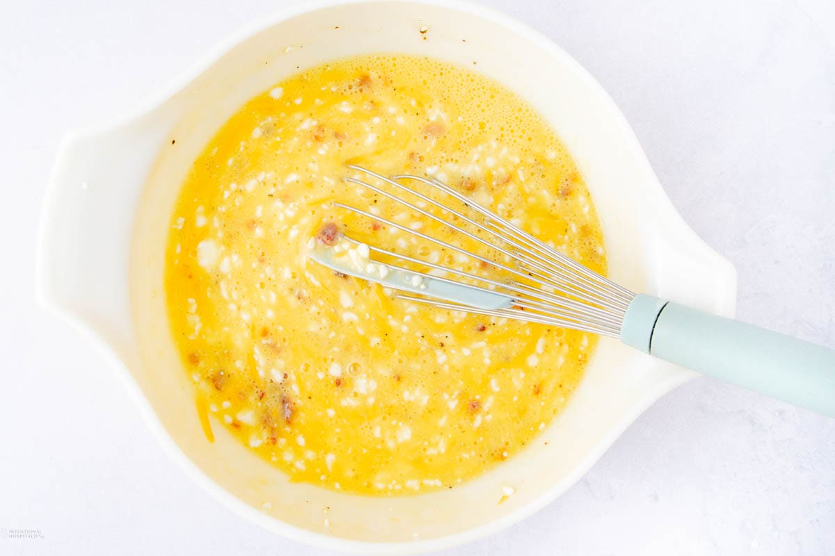 A white mixing bowl contains a beaten egg mixture with chunks, likely cheese or ham, and a few black pepper specks. A whisk with a light blue handle rests inside, suggesting the start of cottage cheese egg bites preparation.