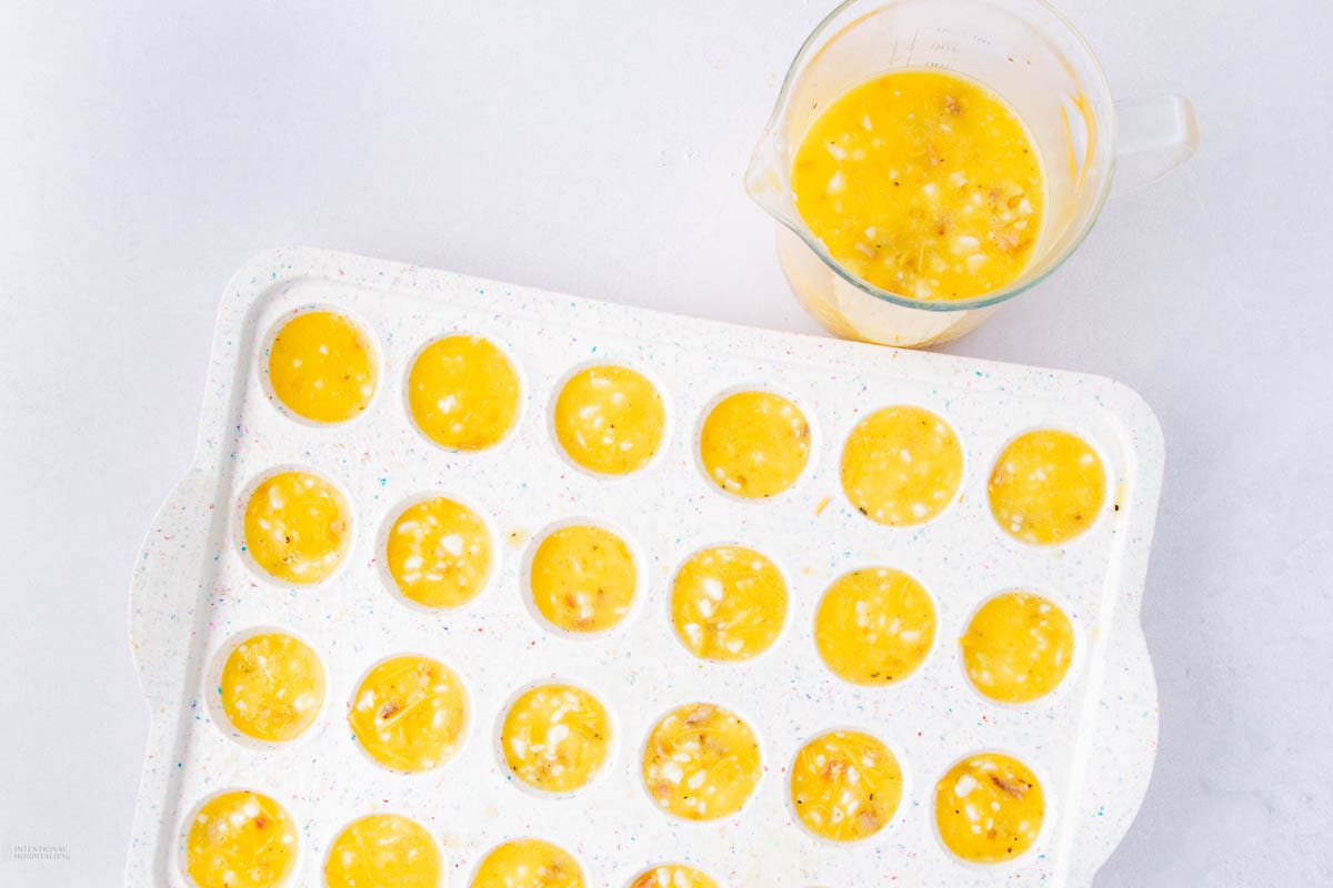 A white silicone mold filled with a yellow egg mixture, likely blended with cottage cheese and bits of onion or vegetable, sits beside a measuring cup containing more of the same. The scene is set on a pristine white surface.