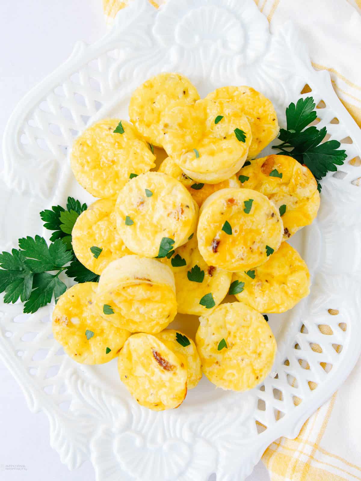 A white plate filled with small, round cottage cheese egg bites garnished with chopped parsley. The plate rests on a patterned cloth, and some parsley sprigs are on the side for decoration.