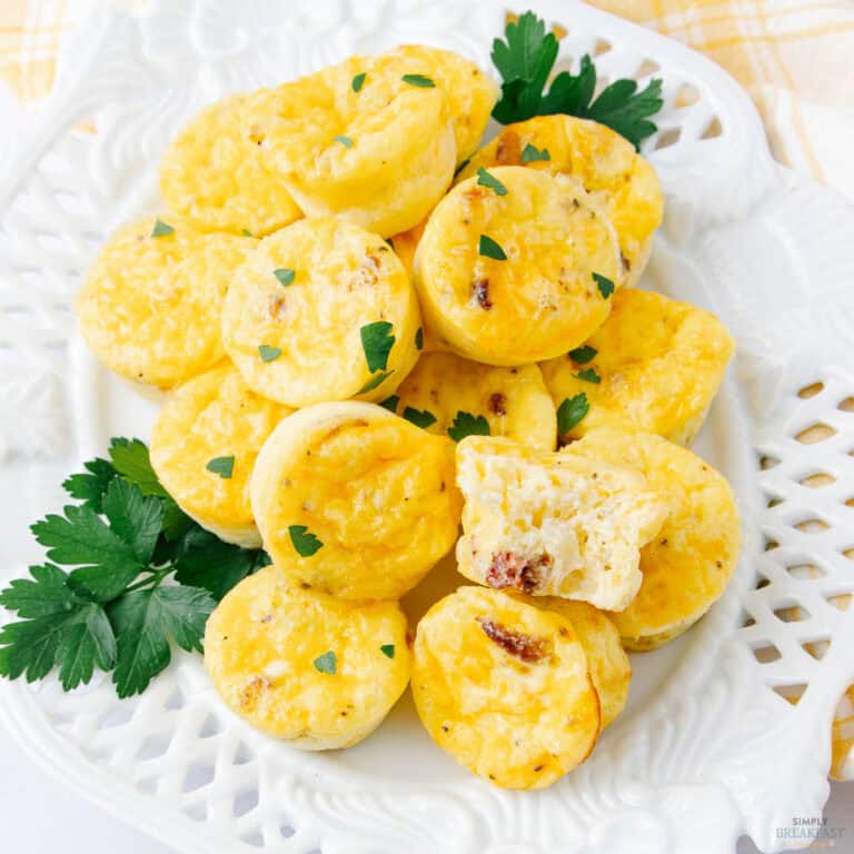 A white plate filled with small, round yellow cottage cheese egg bites garnished with parsley. One bite is cut open, revealing a soft, creamy texture inside. Fresh parsley leaves are placed around the plate for decoration.