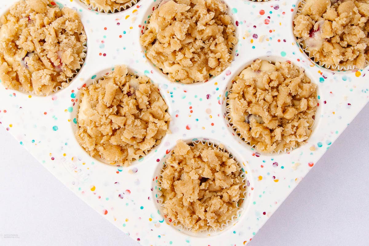 A baking pan filled with unbaked muffin cups, each topped with a crumbly streusel mixture. The pan is white with colorful speckles, adding a festive touch to the scene.