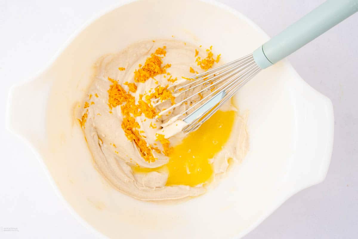 A white mixing bowl contains partially mixed batter with orange zest and egg yolk, alongside a light blue whisk resting inside. The ingredients are ready to be blended further.