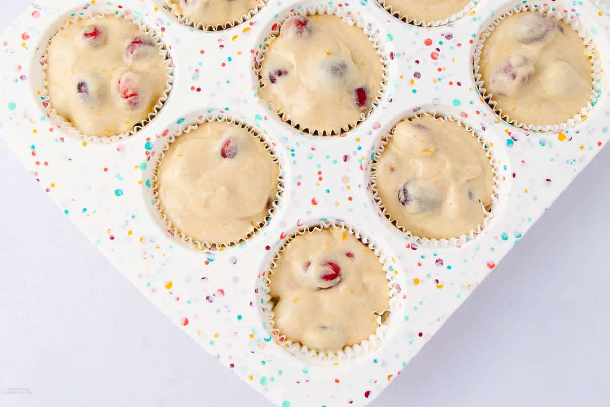 A muffin tin filled with batter-stuffed paper liners, each containing visible red and dark berries. The tin features a colorful, speckled pattern on a white background.