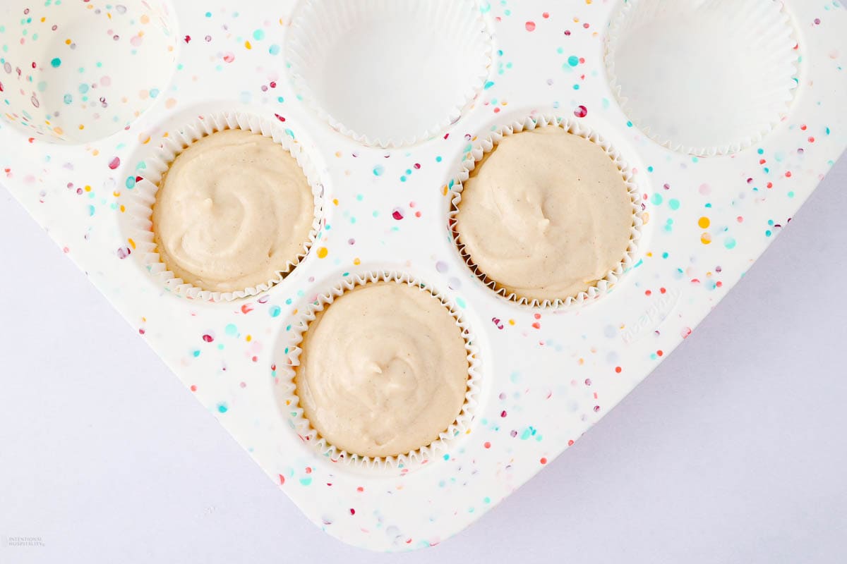 Muffin tray with three filled cupcake liners showcasing creamy batter, surrounded by three empty liners. The tray features colorful confetti-like dots on a white background.