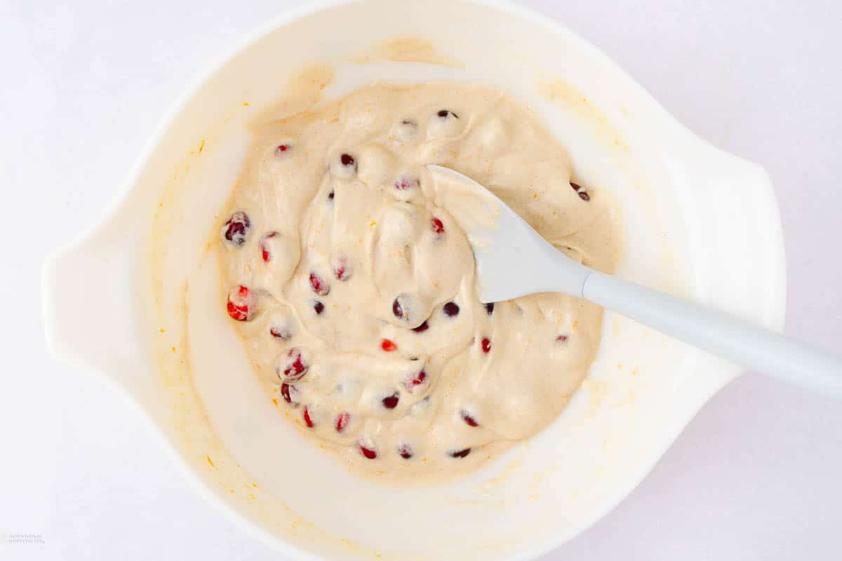 A mixing bowl contains creamy batter with visible cranberries. A spatula rests inside, positioned as if stirring. The background is a neutral surface, emphasizing the bowl's contents.