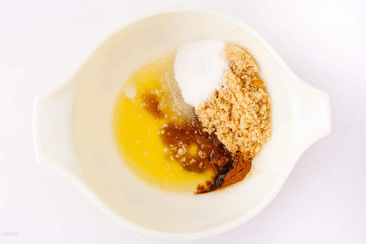 A mixing bowl with melted butter, brown sugar, white sugar, and cinnamon. The ingredients are placed separately before mixing, on a white background.