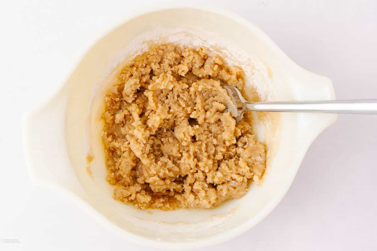 A white mixing bowl containing brown, crumbly cookie dough with a spoon. The bowl is on a white surface.