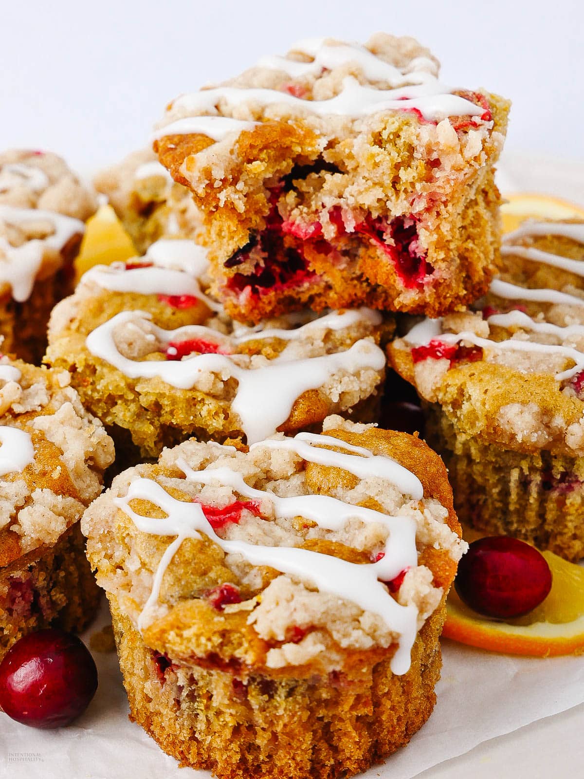 Close-up of a stack of cranberry muffins topped with a white glaze. The muffins are moist, with visible cranberries and crumbly streusel. The background includes orange slices and whole cranberries for decoration.