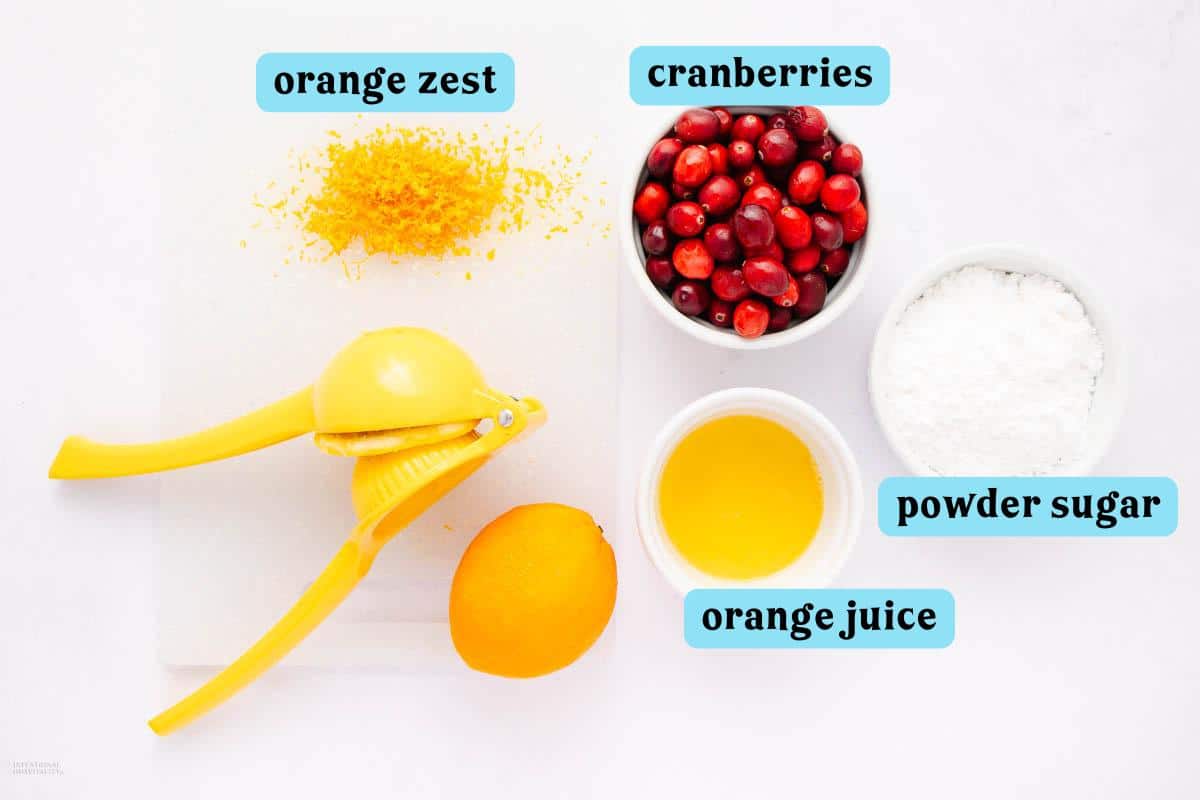 A lemon squeezer, an orange cut in half, orange zest, cranberries in a bowl, orange juice in a cup, and a bowl of powdered sugar on a white surface. Labels indicate each item: "orange zest," "cranberries," "powder sugar," and "orange juice.