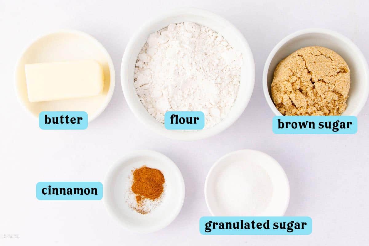 Top view of six bowls containing butter, flour, brown sugar, cinnamon, granulated sugar, and a pinch of cinnamon, labeled in blue text. The ingredients are arranged on a white background.