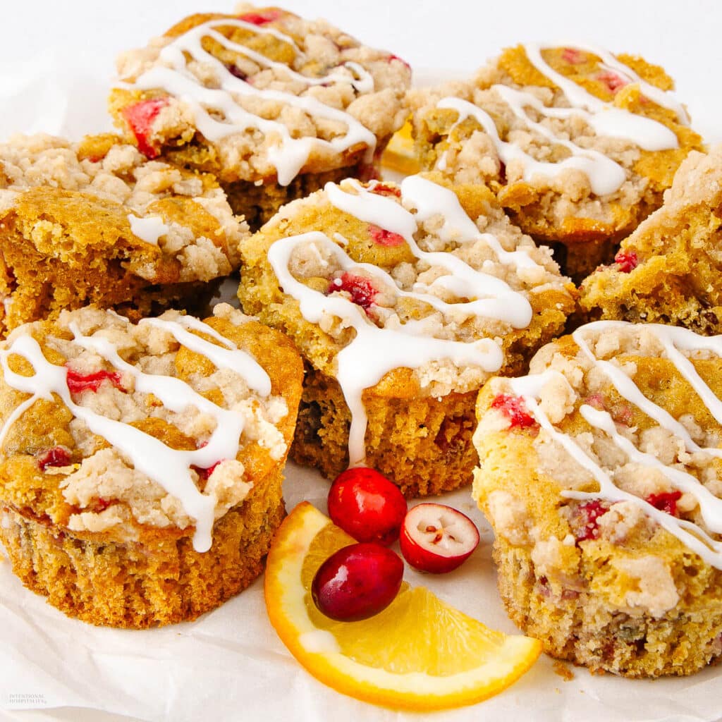 Freshly baked cranberry orange muffins with crumb topping and white icing drizzled on top. The muffins are arranged on a white surface with scattered cranberries and an orange slice.