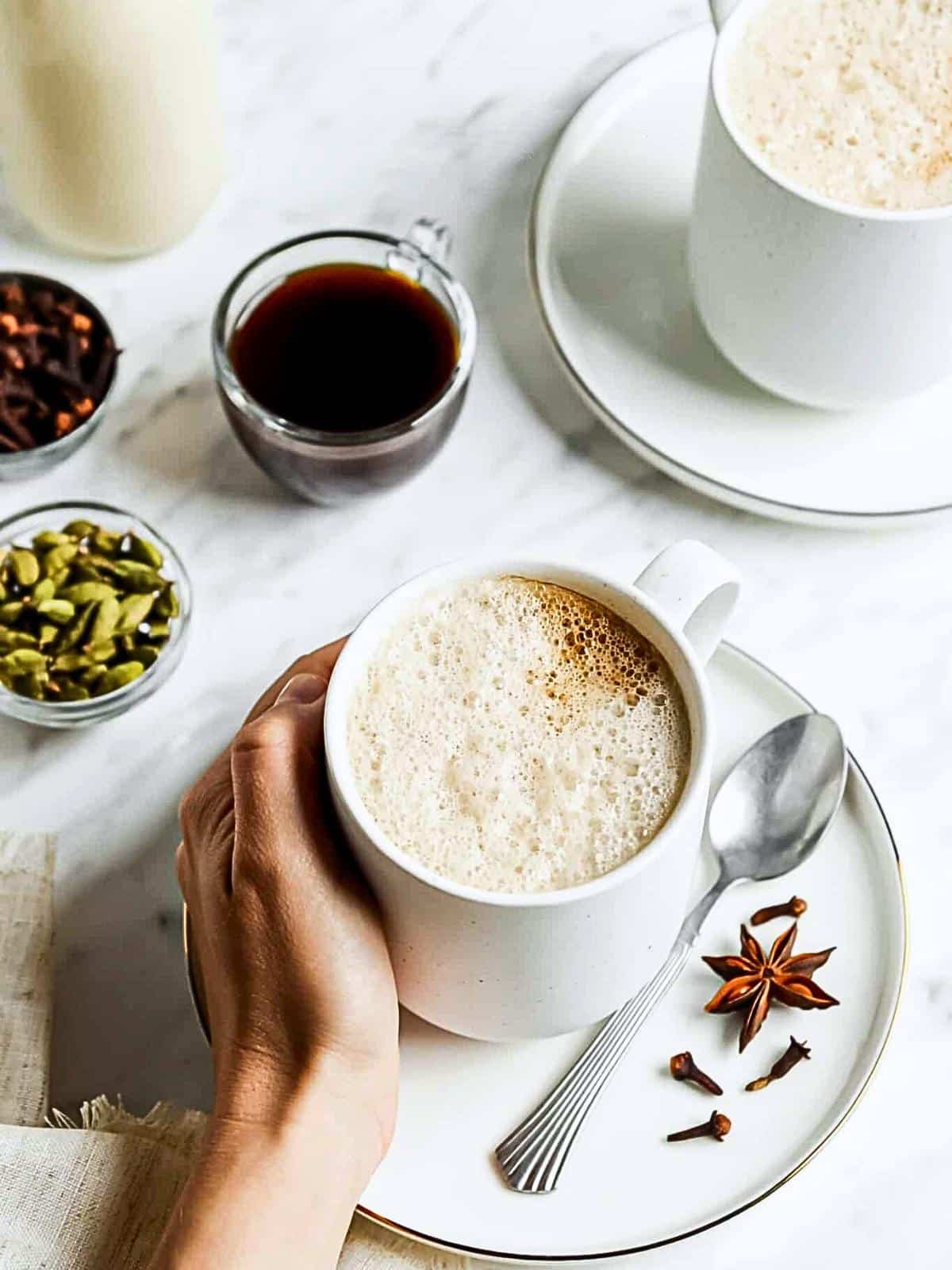 A hand holds a white mug of frothy coffee on a table. Nearby are a spoon, star anise, a small bowl of cardamom pods, a cup of black coffee, and another white mug on a white saucer. A light, airy setting with a focus on warm beverages and spices.