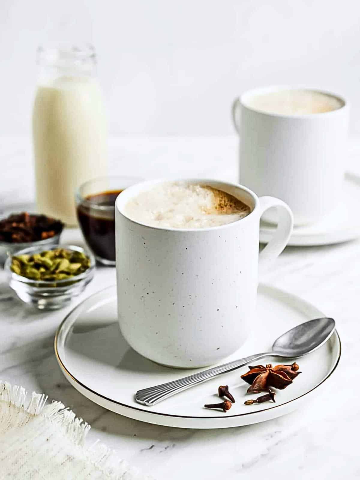 A white ceramic mug filled with frothy chai sits on a saucer with a teaspoon. Star anise and cloves are scattered on the saucer. In the background, there's another mug, a bottle of milk, and bowls of spices. The scene is set on a marble surface.