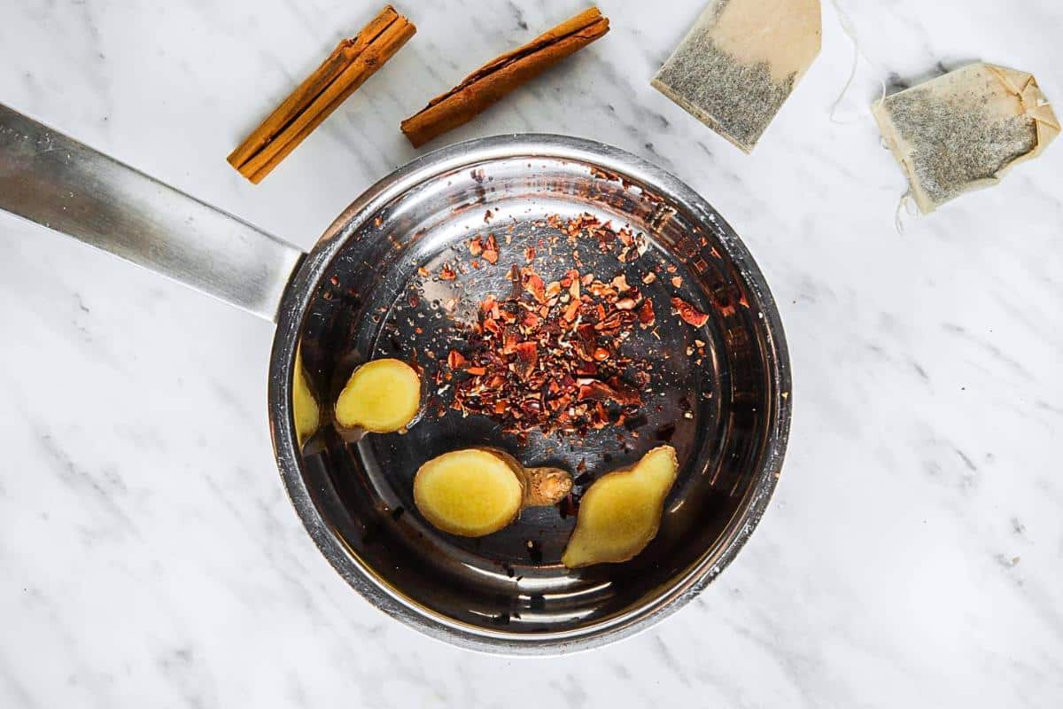 A metal pot on a marble surface contains sliced ginger, cinnamon sticks, and tea leaves. Two tea bags rest nearby.